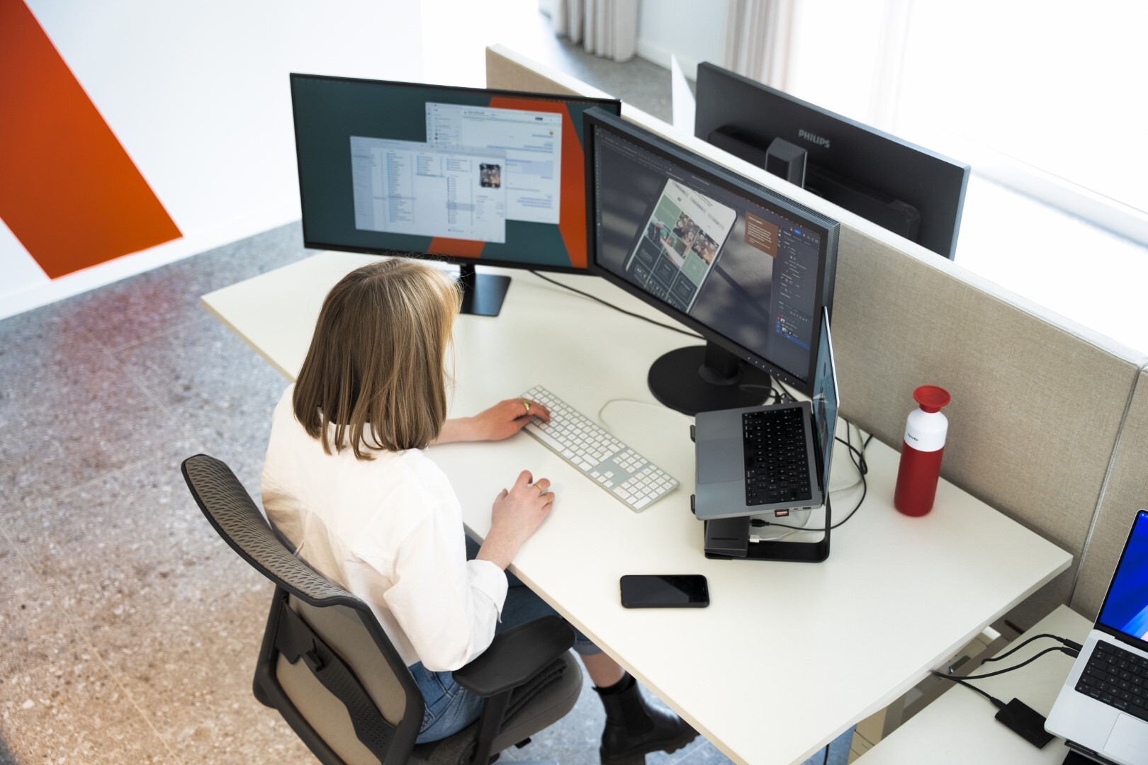 Een vrouw die zit aan een bureau met twee schermen en een laptop