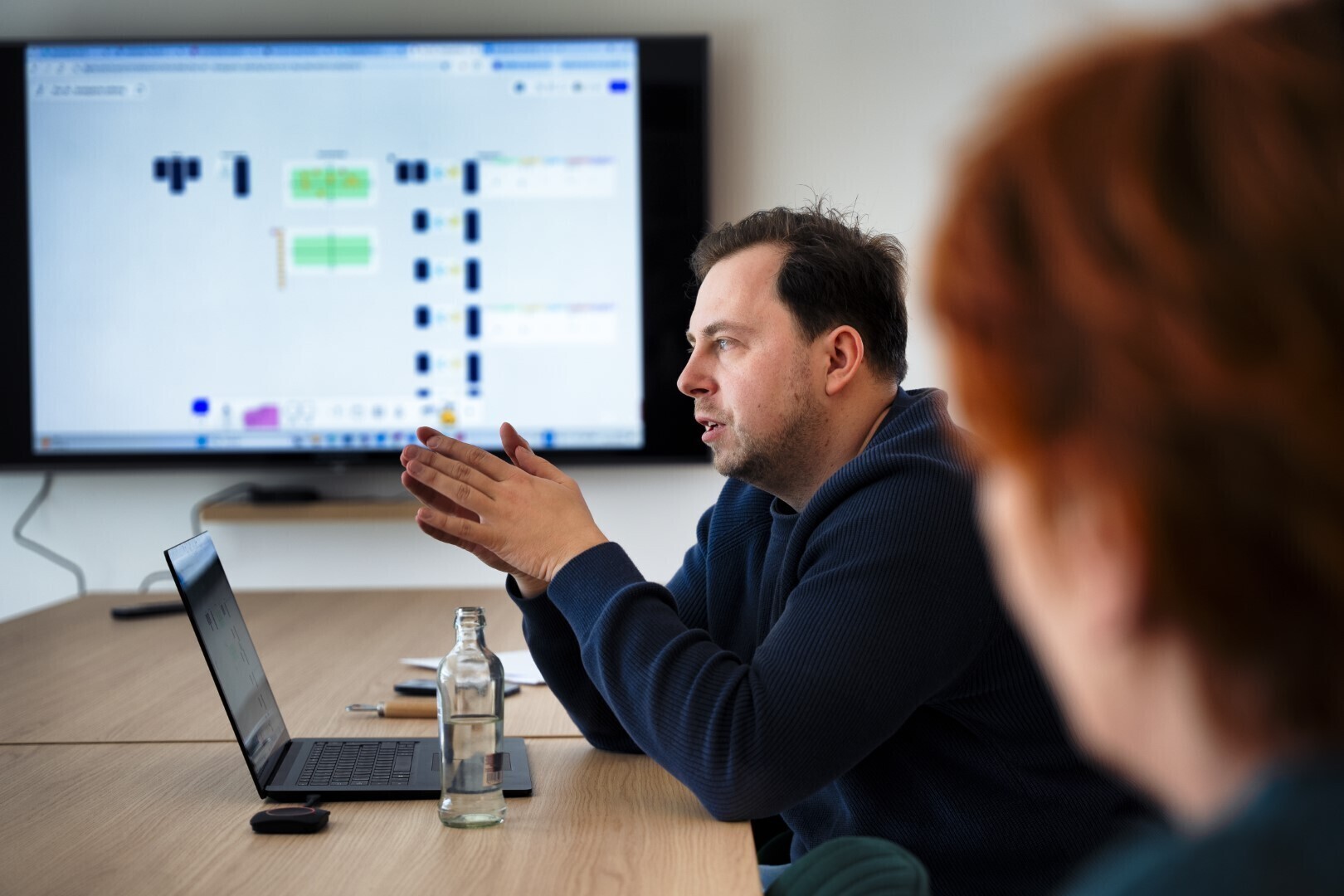 Man zit aan een vergadertafel met een laptop en spreekt terwijl er een scherm met een workflow of planning op de achtergrond hangt.