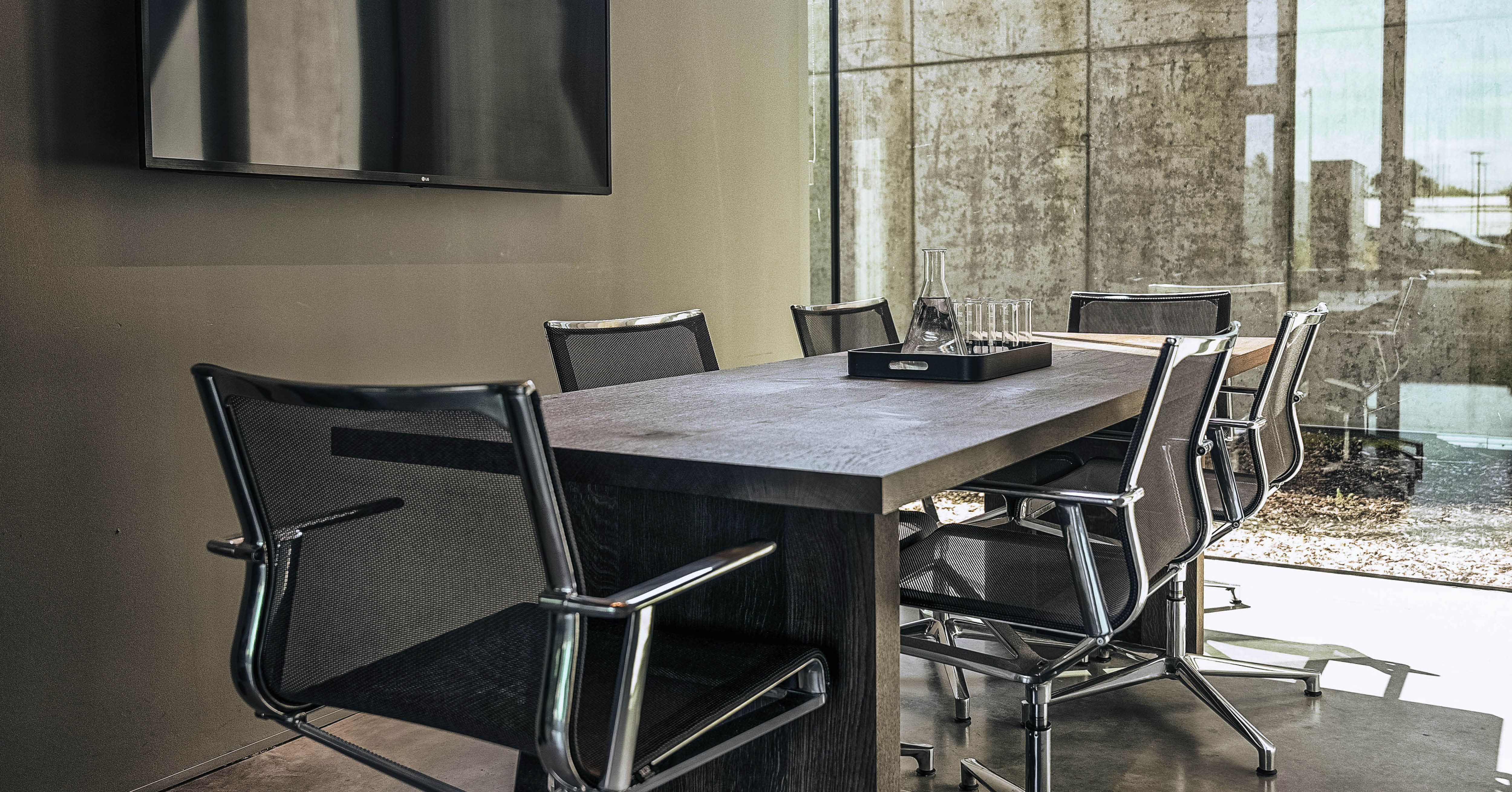Vergaderruimte met zwarte tafel, metalen stoelen en groot raam dat natuurlijk licht binnenlaat, met een flatscreen aan de muur.