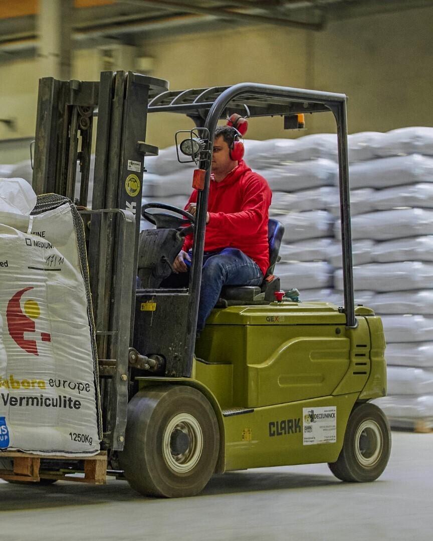 Man in rode jas bestuurt een heftruck met een bigbag in een magazijn, omringd door stapels witte zakken.
