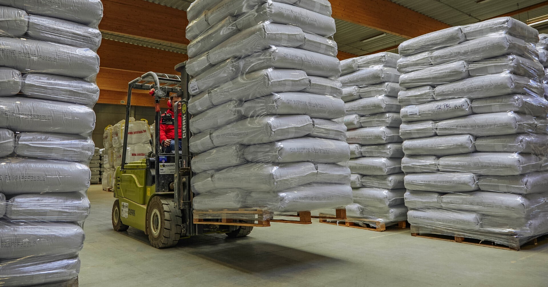 Heftruckchauffeur verplaatst een hoge stapel verpakte zakken op een pallet in een ruim magazijn met meerdere stapels zakken rondom.