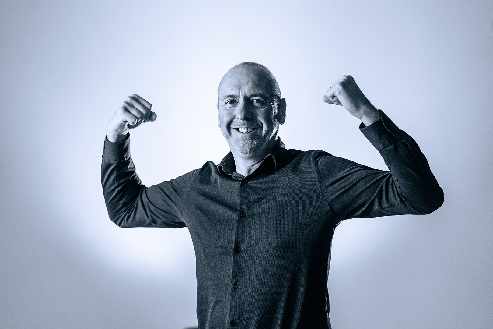 Glimlachende kale man in zwarte blouse toont spierballen met opgeheven armen, zwart wit foto