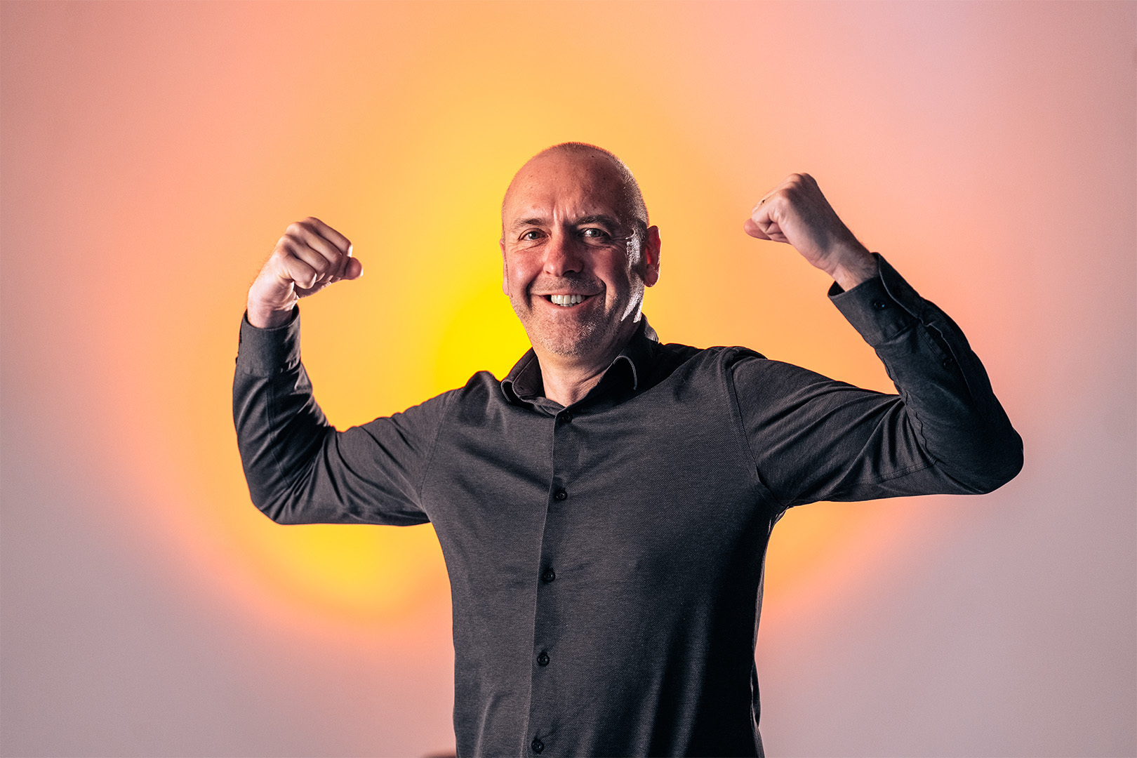 Glimlachende kale man in zwarte blouse toont spierballen met opgeheven armen, zwart wit foto