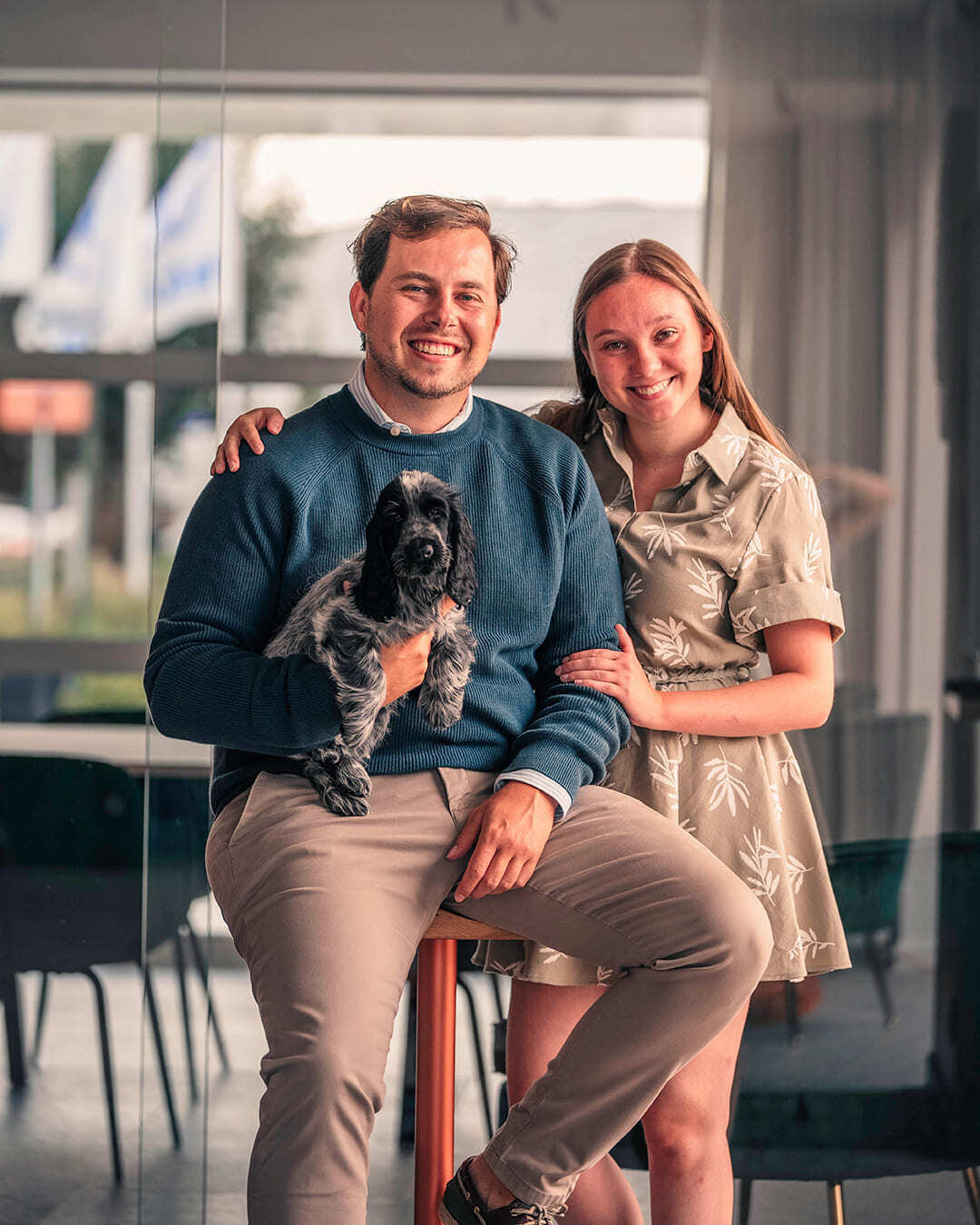 Een koppel kijkt lachend in de camera, mekaar vasthoudend en hun hondje op de arm