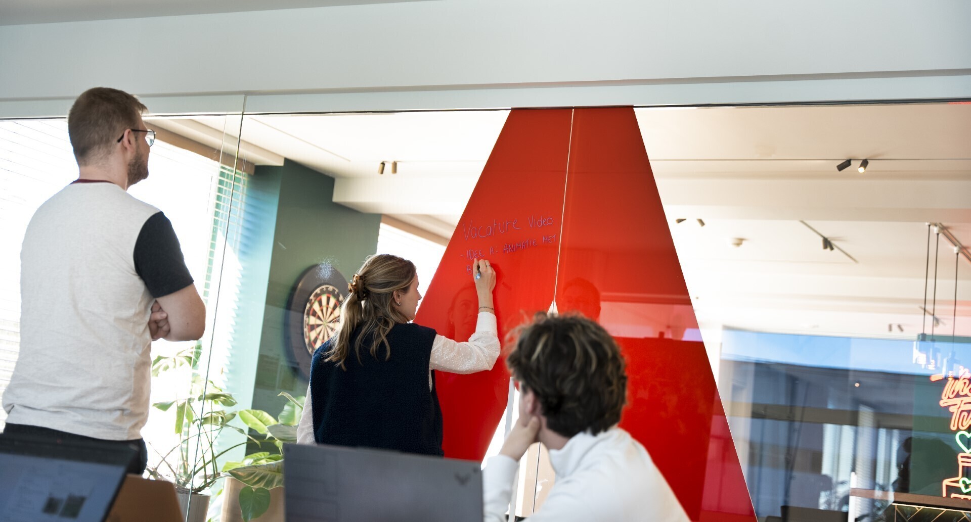 Teamleden brainstormen samen aan een glazen muur in een creatief kantoor, met een opvallend rood element op de achtergrond.